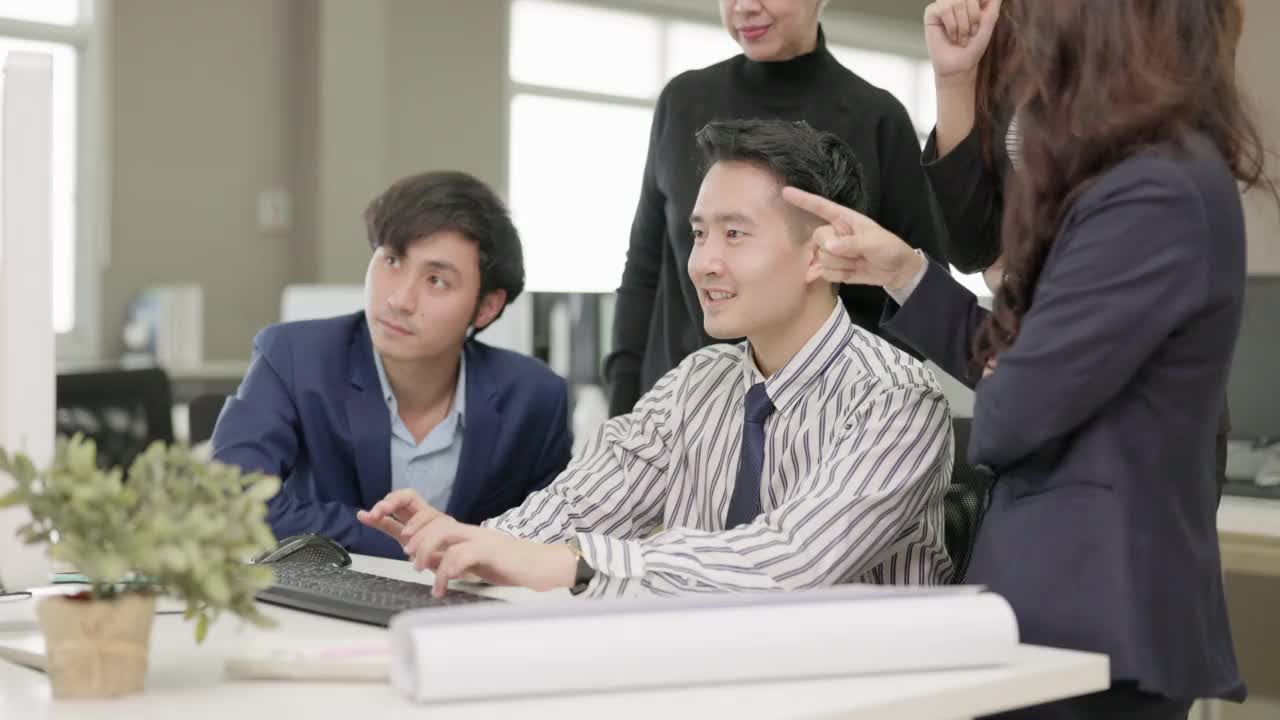 戴眼镜的办公室男性在电脑上听到好消息时，与同事们拥抱并挥手庆祝。