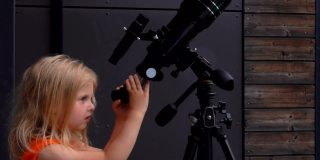 一个金发小女孩在橘黄色的顶部正在通过望远镜探索天空