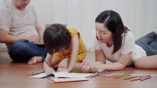 亚洲幸福家庭在客厅一起做活动。妈妈教孩子画画，她和孩子们一起躺在地板上。家庭关系视频素材模板下载