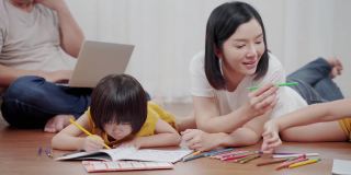 亚洲幸福家庭在客厅一起做活动。妈妈教孩子画画，她和孩子们一起躺在地板上。家庭关系