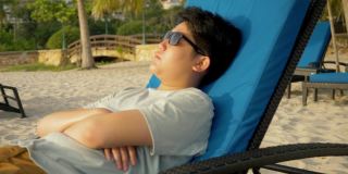 快乐的亚洲男孩戴着太阳镜在沙滩椅上日光浴在早上度假期间在酒店或度假村与家人，生活方式的概念。