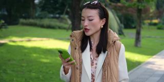 生意小姐下班后走在街上，拿着手机高兴地唱着歌。美丽的中国亚洲女人在户外有视频通话。温暖阳光明媚的秋日。