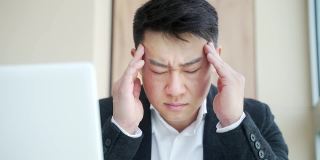 年轻的亚洲男子，办公室职员坐在工作场所握着手按摩额头和头部与严重的疼痛