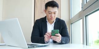 年轻的亚洲上班族坐在办公室里，用智能手机聊天发短信