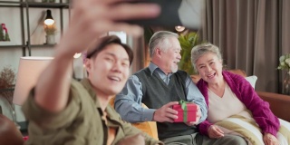 笑容满面的亚洲大家庭和孩子们坐在客厅的沙发上一起在手机上自拍视频，快乐的父母和孩子们在家里用智能手机进行有趣的视频通话