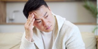 一个年轻的亚洲男人在家里的沙发上抱着他严重头痛的头