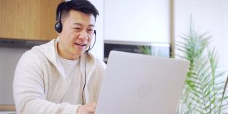 年轻的亚洲人自由职业者坐在家里的办公桌与耳机和通过笔记本电脑在房间或厨房在线视频连接