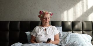 早上醒来，50多岁的女人拿着卷发器坐在床上看电视，用遥控器换频道