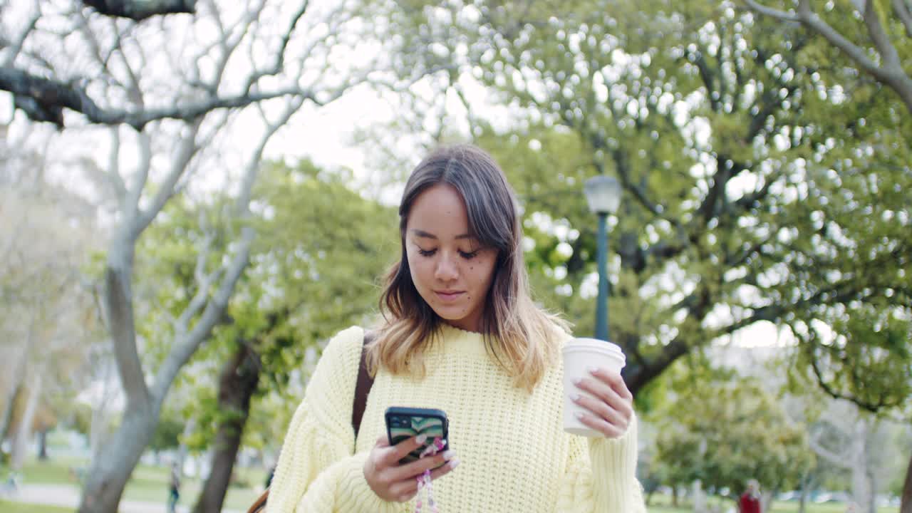 一段4k视频记录了一位迷人的年轻女子边喝咖啡边发短信边走过公园