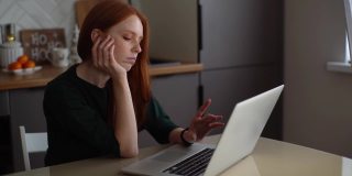 疲惫无聊的年轻女子在笔记本电脑键盘上打字，手放在下巴坐在桌子上的厨房室内现代。