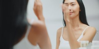 美丽的亚洲女人触摸她完美柔软的皮肤和繁茂的黑发，微笑在浴室的镜子。健康健康的概念，自然美容，有机护肤品