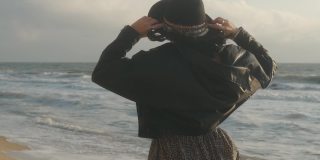 在秋天的日落或日出时，浪漫的黑发女人戴着帽子和皮夹克在海滩上散步