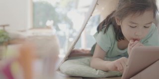 混合亚洲女孩远程学习，用数码平板电脑视频通话在一个再利用的纸板帐篷在家里，使用缩放在线虚拟课堂，家庭教育，新常态概念