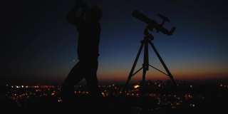 星空下一个男人的剪影，望远镜和城市街灯。