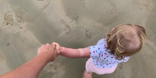 蹒跚学步的小女孩在海滩上牵着妈妈的手，从山顶往下看