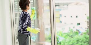 亚洲男孩戴防护手套，通过喷洒清洁产品和用海绵擦拭窗户。家庭家务与家庭观念。