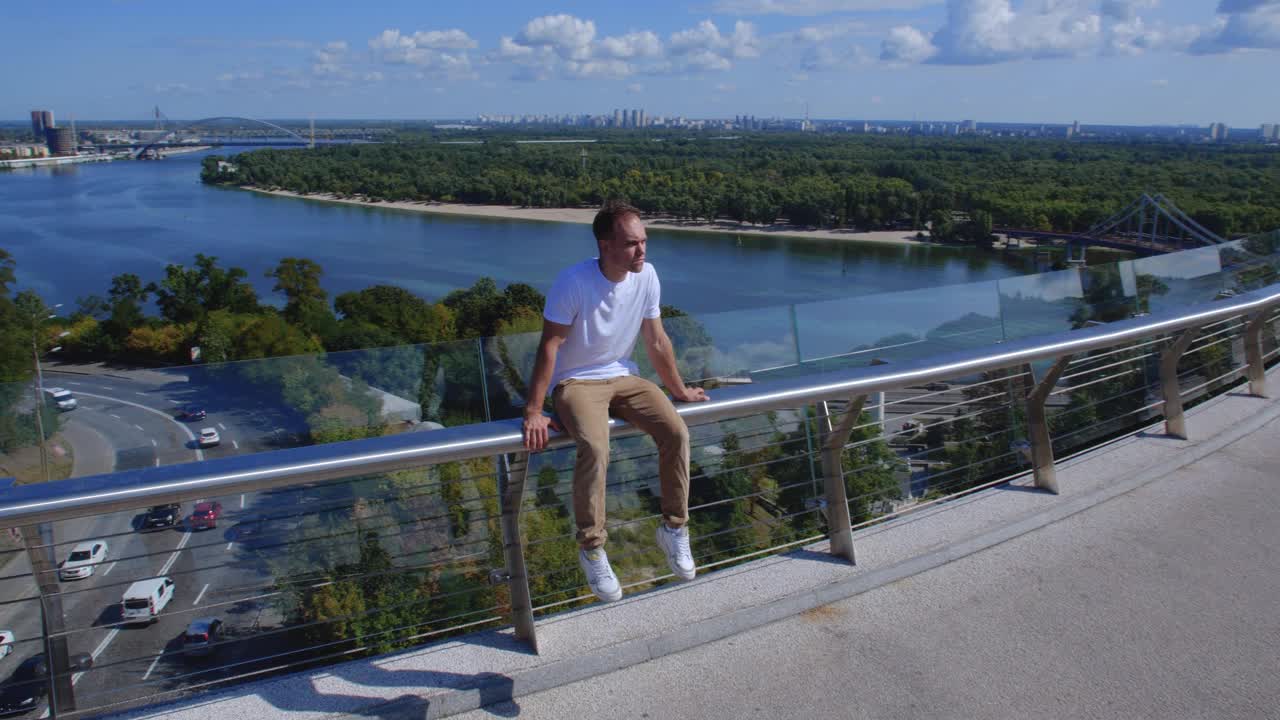 跟踪拍摄一个人坐在玻璃桥的栏杆上，以城市为背景。一个白人坐在一根狭窄的金属管上。失去希望和自杀的概念。总会有出路的