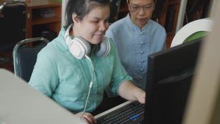 真实的盲人女性带着耳机使用电脑和盲文显示辅助装置与高级同事在工作场所讨论，手持拍摄。视频素材模板下载