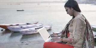 一个年轻漂亮的女人穿着传统的沙瓦卡米兹，坐在河边的平台上用笔记本电脑工作，