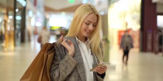 微笑优雅的年轻女子的肖像与长长的金发看着相机和使用手机站在购物中心的纸袋。