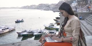 一个年轻漂亮的女人穿着传统的沙瓦卡米兹，坐在河边的平台上用笔记本电脑工作，