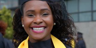 美丽的非裔美国女大学毕业生穿着毕业礼服和学位帽对着镜头微笑着露出牙齿
