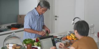 亚洲华人老人在厨房柜台准备食物，切蔬菜，妻子用笔记本电脑回复电子邮件，享受亲密时光
