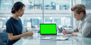 多民族多元化办公会议室会议:两位创意企业家团队讨论成长策略，使用带绿屏的笔记本电脑模拟显示器。
