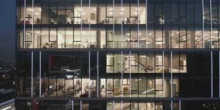 鸟瞰图的现代办公室在玻璃摩天楼在晚上，全景左边。商务人士在办公室里对着电脑工作到很晚。办公楼夜间灯火通明。