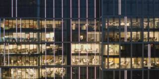 鸟瞰图的现代办公室在玻璃摩天楼在晚上，全景左边。商务人士在办公室里对着电脑工作到很晚。办公楼夜间灯火通明。