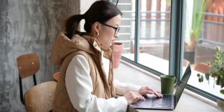 漂亮的亚洲华人女商人戴着眼镜，在咖啡馆靠窗的地方用笔记本电脑做自由职业者。休息时间上网工作。敲击键盘。年轻的学生在咖啡馆学习