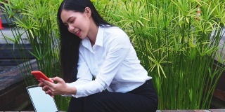 快乐的年轻亚洲妇女与笔记本电脑使用她的智能手机通过wi-fi互联网在公园交流。