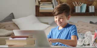 微笑的学生男孩在家学习与远程学校连接到网络上的电脑，举起他的手回答问题或问候他的同学，坐在堆满了书的书桌
