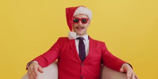 一个快乐的小胡子男人，穿着红色的衣服，戴着圣诞老人帽，戴着红色滑稽的眼镜，对着相机微笑，在黄色的背景上捕捉圣诞礼物。新年销售和促销概念