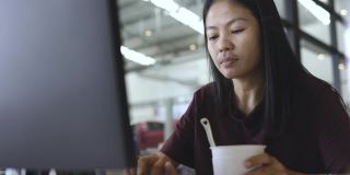 亚洲自由职业者妇女吃方便面杯和工作在电脑屏幕前