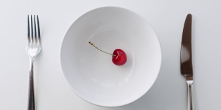 一个多汁的红樱桃从高处落在一个孤立的空盘上，刀叉放在白色背景的餐桌上。桌面慢动作拍摄，饮食极简主义午餐理念
