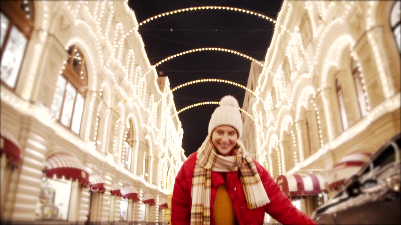 一个美丽快乐的女孩，穿着红色的夹克，戴着白色的帽子和围巾，在灯火通明的圣诞街上跑着，微笑着调情。户外城市快乐新年圣诞时间概念