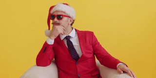 悲伤的小胡子男人穿着红色的西装，戴着圣诞老人的帽子，戴着红色滑稽的眼镜，坐在椅子上，收到了一个惊喜的礼物，感到非常高兴。新年销售和促销惊喜哇概念