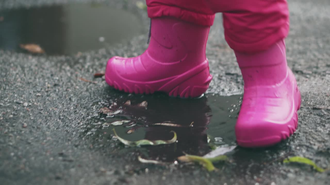 快乐的小女孩跑过水坑，特写。可爱的小女孩在雨后穿着粉红色的胶靴走路。