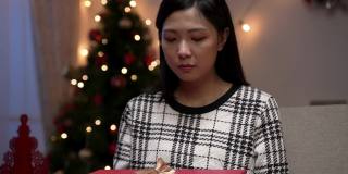 快乐的亚洲女性捂住她的嘴与惊讶的脸，当她打开和看到美丽的圣诞礼物在一个舒适的客厅在晚上