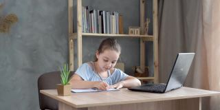 可爱的十几岁女学生在家里用笔记本电脑做作业。孩子把任务记在笔记本上。
