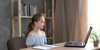 小白人女孩微笑和挥手，而坐在家里的桌子上与笔记本电脑。在线交流，远程学习。