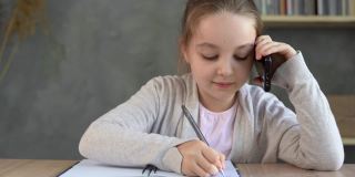 一个女学生坐在家里的桌子边打电话，在笔记本上做笔记。