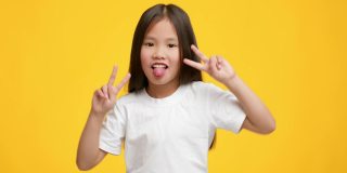 亚洲小女孩胜利的手势，伸出舌头，黄色背景
