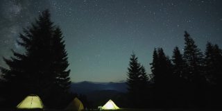 在漆黑的群山中，在星光闪烁的夜空下，徒步旅行者在明亮的旅游帐篷旁的篝火旁休息。积极的生活方式和户外生活理念