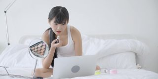 年轻的亚洲女性美女在床上用口红学习化妆和在线教程看着笔记本电脑，女性在流媒体上教授和讲解化妆和化妆。