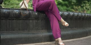 一位无法辨认的女子坐在城市公园的喷泉旁，双腿交叉在膝盖上，慢动作。苗条时尚的白种人女士穿着紫色连衣裤和高跟鞋在夏日户外休息