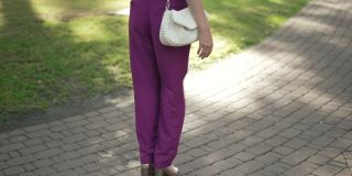 女性的双腿漫步在阳光明媚的夏日公园户外的人行道上。无法辨认的白人苗条优雅的女人在阳光下慢动作穿着紫色连衣裤和高跟鞋行走