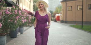一个积极的女人穿着紫色的连衣裤，戴着草帽，慢动作地走在镇上的户外四处张望。前视图肖像自信优雅苗条美丽的成年女士漫步在城市的早晨