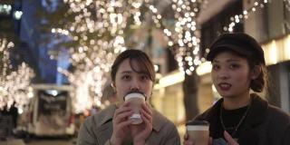 年轻的女性朋友在圣诞的灯光下在城市的街道上喝咖啡